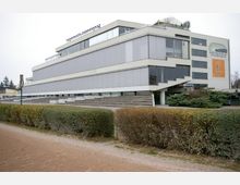 Modernes Gebäude mit flacher, rechteckiger Bauweise, bestehend aus mehreren Etagen mit großen, teils geschlossenen Glasflächen und einem Treppenaufgang. Im Vordergrund befinden sich ein Gehweg sowie gepflegte Hecken.