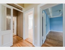 Innenansicht eines Wohnungsflurs mit hellen Holzdielen, hohen weißen Türzargen und mehreren offenen Durchgängen zu angrenzenden Räumen. Rechts ist ein blau gestrichenes Zimmer mit Stuck an der Decke und Deckenleuchte sichtbar, links eine Tür mit Glasfeld und dekorativem Muster.