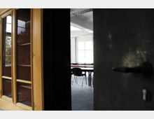 Ein teilweise geöffneter Raum mit dunkler Tür im Vordergrund, dahinter ein heller Besprechungsraum mit Tischen, Stühlen und großen Fenstern, die Tageslicht hereinlassen. An der linken Seite steht ein Holzschrank mit Glastüren.