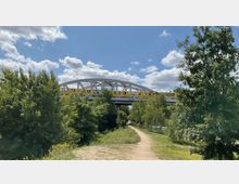 Ein gelber Zug fährt über eine graue Stahlbrücke, die von Bäumen und Grünflächen umgeben ist. Vorne verläuft ein sandiger Fußweg durch die Landschaft, während darüber ein blauer Himmel mit Wolken zu sehen ist.