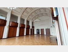 Ein großer, hoher Raum mit gewölbter Decke, Holztüren und Emporen aus dunklem Holz. Der Boden besteht aus hellem Parkett, und eine runde Deckenleuchte mit kugelförmigen Lampen hängt zentral im Raum.