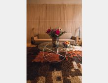 Wohnzimmer mit einer beigefarbenen Couch, einer Holzpaneelwand und einem runden Glastisch mit einem Metallgestell. Auf dem Tisch steht eine Vase mit Blumen und eine Schale mit Früchten, während unten ein flauschiger Teppich in Brauntönen liegt; an den Seiten sind Akustikgitarren zu sehen.