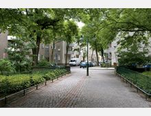 Gepflasterter Gehweg in einem Wohngebiet, gesäumt von grünen Bäumen und Metallzäunen. Im Hintergrund sind mehrstöckige Wohnhäuser sowie parkende Autos zu sehen.