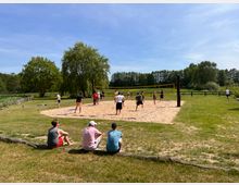 Eine Gruppe von Menschen spielt Volleyball auf einem Sandplatz, umgeben von einer grünen Wiese und Bäumen. Im Vordergrund sitzen drei Zuschauer auf einem Holzstamm, während der Hintergrund eine natürliche Landschaft mit Zaun und Wald zeigt.