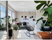 Helles Wohnzimmer mit modernen Möbeln, darunter ein Schaukelstuhl, ein Holztisch und ein Esstisch mit schwarzen Stühlen. Große Fenster lassen viel Tageslicht herein; im Raum stehen zahlreiche grüne Zimmerpflanzen. Im Hintergrund ist eine kleine Küchenzeile und ein gerahmtes Foto an der Wand erkennbar.