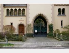 Historische Gebäudeansicht mit einem großen Spitzbogenportal und einem verzierten Metallgittertor. Links daneben befindet sich eine Holztür mit einem dekorativen Rundbogen, umgeben von gepflegten Büschen und einer kleinen gepflasterten Fläche.