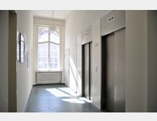 Ein heller Flur in einem Gebäude mit zwei Aufzugtüren auf der rechten Seite, einem großen, bogenförmigen Fenster im Hintergrund, das Tageslicht hereinlässt, und einem Heizkörper unter dem Fenster. Links sind Bilder und Aushänge an der Wand zu sehen.