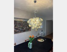 Ein modernes Esszimmer mit einem schwarzen Tisch, auf dem eine Vase mit roten und gelben Tulpen sowie Teller mit Snacks stehen. An der Decke hängt eine dekorative Hängelampe aus vielen glänzenden Glaskugeln, im Hintergrund ist ein farbiges Wandgemälde zu sehen.