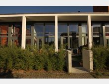 Moderne Wohnhausfassade mit großen Glasfenstern und weißen Betonstützen. Im Vordergrund befinden sich eine niedrige Hecke und ein gepflasterter Zugang, der von zwei Betonpfeilern mit der Nummer „12E“ eingerahmt ist.