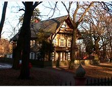 Historisches Fachwerkhaus mit Spitzdach, umgeben von hohen Bäumen in einer herbstlichen Parklandschaft. Im Vordergrund ist ein Zaun mit ornamentaler Verzierung zu sehen.