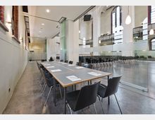 Moderner Seminarraum in einer umgebauten Kirche, mit hohen Decken, großen Rundbogenfenstern und einer Glaswand, die den Besprechungsbereich mit Tisch und Stühlen von einem größeren Veranstaltungsraum trennt. Der Boden ist aus Beton, und ringsum hängen schlichte Wandleuchten.