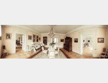 Panoramatische Ansicht eines eleganten, historischen Salons mit pastellfarbenen Wänden und Stuckverzierungen. Der Raum ist mit antiken Möbeln, einem großen Kronleuchter, Gemälden an den Wänden, sowie Teppichen und Weißgold-Verzierungen ausgestattet; zwei offene Türdurchgänge führen zu angrenzenden Räumen.