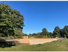 Offene Sandvolleyballplätze mit gelben Netzen in einem Park, umgeben von grünen Bäumen unter klarem, blauem Himmel. Im Vordergrund ist ein Rasenbereich, und orangefarbene Absperrzäune trennen die Sandflächen vom restlichen Gelände.