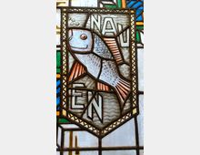 Buntes Glasfenster mit einem Fischmotiv in einem geometrischen Rahmen, umgeben von farbigen Glasstücken in Blau, Grün, Gelb und Weiß. Über und unter dem Fisch sind die Buchstaben „NAU“ und „EN“ in den Rahmen integriert.