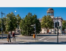 Straßenszene vor einem historischen Gebäude mit Säulen und einer Kuppel, umgeben von Bäumen. Im Vordergrund sind zwei gehende Personen, eine Straßenkreuzung mit Ampeln und ein Straßenschild mit der Aufschrift „Schloßstraße“ sichtbar.