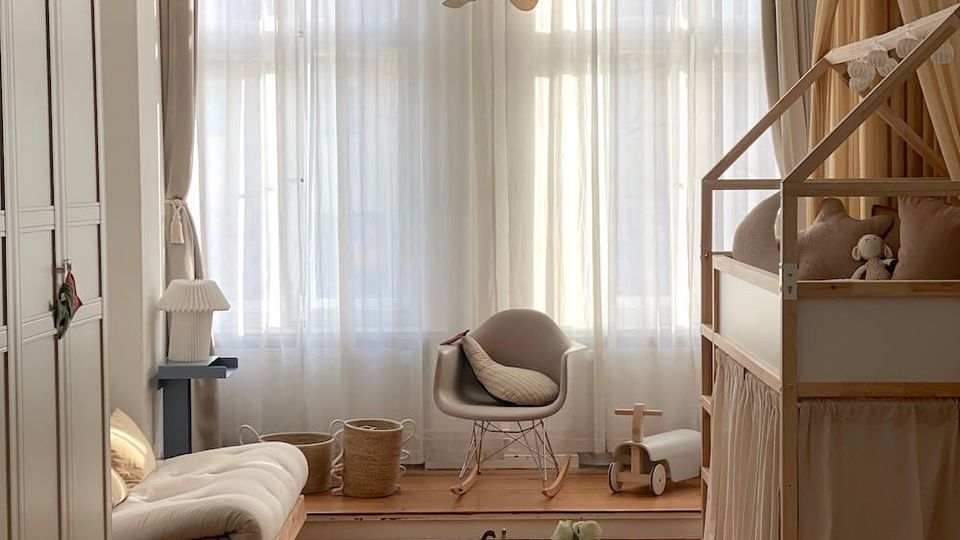Bright children’s bedroom with large curtained windows, a round paper pendant light, and a low table with two small chairs on a circular rug. The room includes a house-frame bed with canopy curtains, a floor mattress, a rocking chair, and light wood flooring with neutral furnishings.
