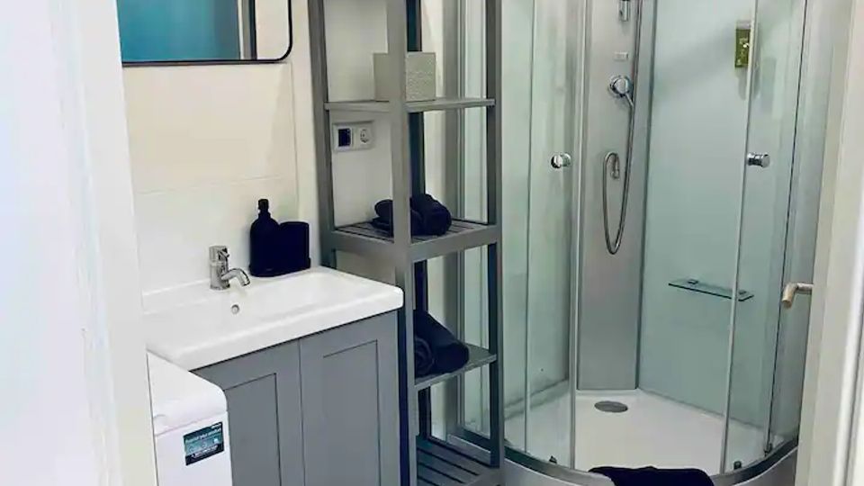 Kleines Badezimmer mit weiß gefliesten Wänden und Bodenfliesen im Rastermuster, links ein Waschbecken mit Unterschrank und Wandspiegel sowie ein schmales Regal. Rechts befindet sich eine abgerundete Glas-Duschkabine mit Handbrause; auf dem Boden liegt ein dunkler Badvorleger.