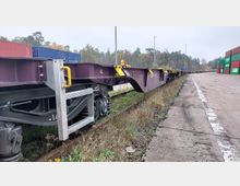 Langer, leerer Güterwagen mit violetter Lackierung und gelben Halteelementen auf einem Bahngleis neben einer asphaltierten Fläche in einem Güterumschlagbereich. Im Hintergrund sind bunte Container sowie Bäume zu sehen.