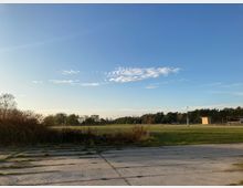 Freifläche mit grünem Rasen und umstehenden Bäumen, im Vordergrund ein Boden aus Betonplatten und niedrige Vegetation; klarer blauer Himmel mit vereinzelten Wolken.