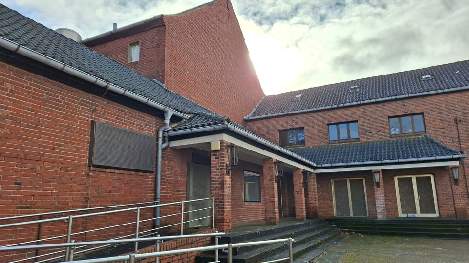 Innenhof eines mehrteiligen Backsteingebäudes mit geneigten Ziegeldächern, Regenrinnen und Wandlampen. Im Vordergrund führen eine Rampe mit Metallgeländer und Stufen zu mehreren Eingängen und großen Türen; darüber ist ein bewölkter Himmel sichtbar.