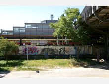 Ansicht eines Stadtbahnhofs mit vorbeifahrendem rot-gelben S-Bahn-Zug, einer mit Graffiti besprühten Wand im Vordergrund und moderner, verglaster Bahnhofarchitektur im Hintergrund. Links und rechts stehen vereinzelte Bäume, während ein Stück einer Stahlkonstruktion am rechten Bildrand sichtbar ist.