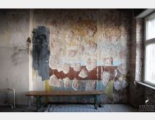 Ein leerer, verwitterter Raum mit einer stark abgenutzten Wand, deren Putz teilweise abgeblättert ist und verschiedene Farbschichten sichtbar macht. Im Vordergrund steht eine einfache Holzbank vor einer gefliesten Bodenfläche, rechts ein Fenster mit Tageslichteinstrahlung.