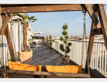 Eine moderne Dachterrasse mit Holzbelag, dekoriert mit Topfpflanzen wie Palmen und Buchsbäumen. Im Vordergrund ist eine Holzschaukel mit orangefarbenen Kissen, und im Hintergrund befinden sich Sitzmöbel, Sonnenschirme und Lichterketten, die die gemütliche Atmosphäre ergänzen.