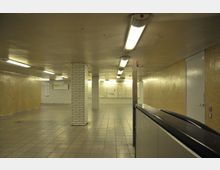 Unterirdischer U-Bahn-Zugang mit gelb verfliesten Wänden und weißgekachelten Säulen. Deckenleuchten beleuchten den Raum, der mit grauen Fliesen ausgelegt ist, und es sind Infotafeln an der Wand sichtbar.