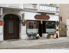 Fassade eines Altstadtcafés mit braunem Holzrahmen und dekorativen Schildern, darauf der Schriftzug „Altstadtcafé Köpenick“. Vor dem Café stehen Blumen in Töpfen sowie ein kleiner Tisch mit zwei Stühlen auf einem gepflasterten Gehweg.