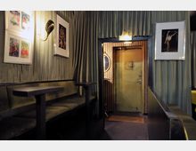 Innenraum einer Bar mit einem Sitzbereich aus gepolsterten Bänken und kleinen Tischen, umgeben von Vorhängen in gedecktem Grün. An den Wänden hängen gerahmte Bilder, und im Hintergrund ist eine Tür mit einem runden Fenster sichtbar, daneben ein Spiegel mit Holzverkleidung.