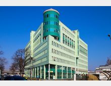 Grünes Bürogebäude mit großen Glasfenstern und einer runden Glasstruktur an der Ecke, gelegen an einer Straße mit Bäumen und mehreren vorbeifahrenden Autos unter klarem blauem Himmel. Im Erdgeschoss befinden sich Geschäfte mit Schildern.