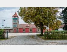Historisches Feuerwehrhaus mit einem hohen Turm und roten Holztoren, umgeben von einem gepflasterten Vorplatz, Bäumen und Hecken. Ein Schild weist auf das Betretungsverbot des Geländes hin.