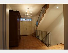 Eingangsbereich mit Terrakotta-Fliesenboden, weißer Doppeltür mit Glaseinsätzen oben, einem großen Holzschrank links und einer Holztreppe mit schwarzem Metallgeländer, die nach oben führt. Von der Decke hängt ein kronleuchterartiger Lampenschirm.