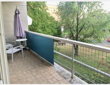 Balkon mit Fliesenboden, weißem Metallgeländer und blauem Sichtschutzpaneel, ausgestattet mit zwei weißen Stühlen, einem kleinen Rundtisch und einem lilafarbenen Sonnenschirm. Im Hintergrund sind Bäume, eine Grünfläche und ein mehrstöckiges Gebäude zu sehen.