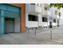 Straßenszene mit einer Wohnhausfassade aus weißem Putz und Balkonen, daneben ein Bereich mit orangefarbenen Kacheln, ein Garagentor und einige Graffiti. Der Gehweg besteht aus Pflastersteinen und Gehwegplatten, im Vordergrund stehen Poller.
