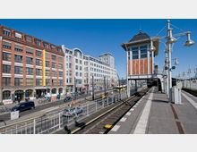 Bahnsteig einer oberirdischen S-Bahn-Station mit einem überdachten Stellwerk aus Holz und Glas rechts im Bild. Im Hintergrund befinden sich mehrstöckige Gebäude im Backstein- und hellen Putzstil entlang einer belebten Straße mit Autos, Straßenbahnschienen und Fußgängern.