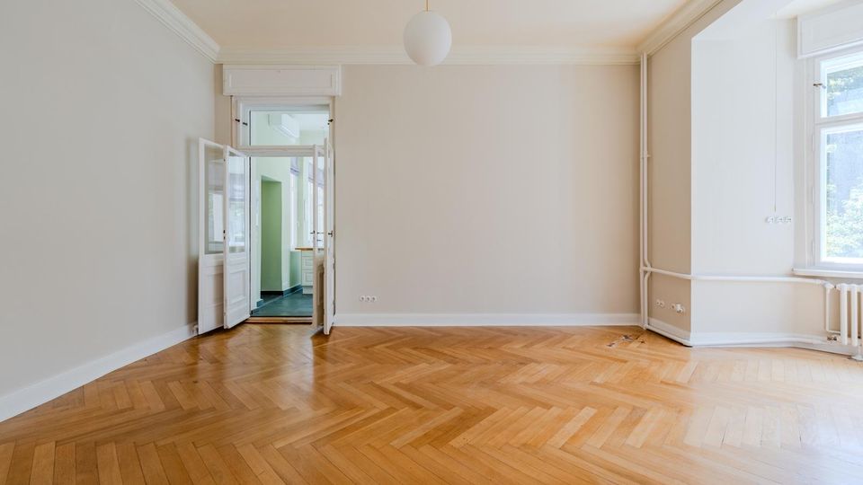 Leerer, heller Wohnraum mit Fischgrät-Parkettboden, weißen Wänden und Stuckleisten an der Decke sowie einer runden Hängelampe. Links führt eine doppelflügelige Tür in einen angrenzenden Raum; rechts sorgen große Fenster, ein Heizkörper und sichtbare Heizungsrohre entlang der Wand für Tageslicht und Details.