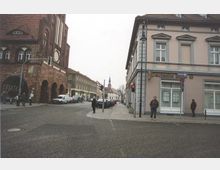 Straßenszene mit historischen Gebäuden: Links ein Backsteingebäude mit Bögen im gotischen Stil, rechts ein helles Gebäude mit klassizistischer Fassade. In der Straße stehen Fußgänger und parkende Autos, im Hintergrund ist der Turm einer Kirche sichtbar.