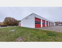 Weißes, einstöckiges Gebäude mit mehreren roten Rolltoren, vermutlich eine Fahrzeughalle, umgeben von Pflasterflächen und Rasen. Im Hintergrund sind Bäume sowie vereinzelte Gebäude sichtbar.