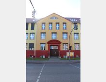 Gelbes und rotes Gebäude einer Musik- und Kunstschule mit symmetrischen weißen Sprossenfenstern, rotem Eingangsportal und einem Vorgarten mit gepflastertem Gehweg. Im Vordergrund eine Straße mit Fußgängerüberweg und einer Ampel.