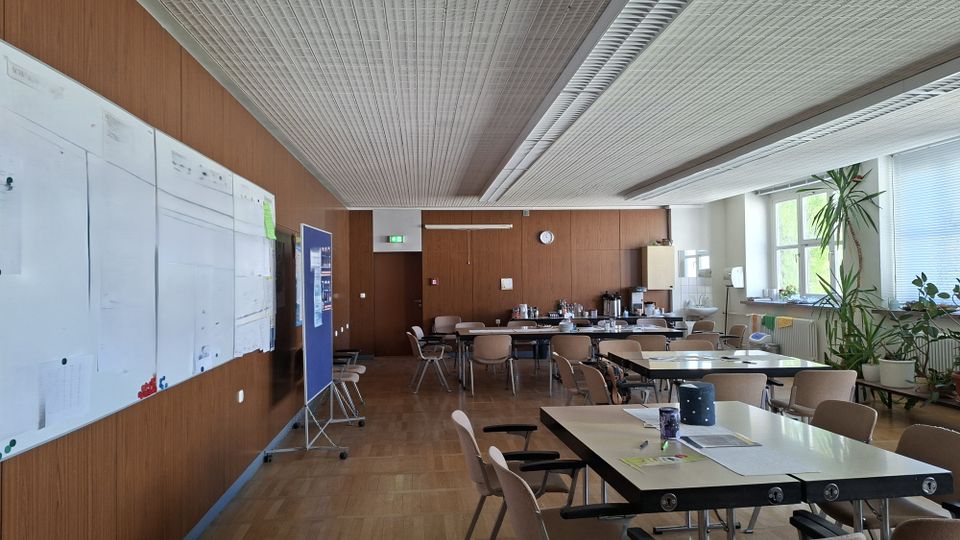 Großer Pausen- oder Aufenthaltsraum mit Holzvertäfelung, Parkettboden und abgehängter Rasterdecke, ausgestattet mit mehreren Tischen und Stühlen. Links hängen große Whiteboards und Stellwände, rechts sorgen große Fenster mit Pflanzen am Rand und eine kleine Küchen- bzw. Getränkezeile im Hintergrund für Tageslicht und Versorgung.