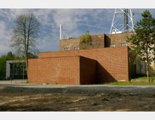 Backsteinbau mit schlichten, rechteckigen Formen, im Vordergrund ein unbefestigter Kiesweg. Im Hintergrund sind weitere Gebäudeteile, ein hoher Sendemast, Bäume und ein klarer Himmel erkennbar.