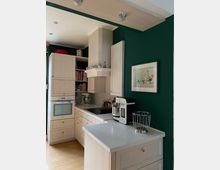 Helle Küche mit weißen Schränken, Einbauherd, Spüle und Arbeitsplatte aus hellem Material. Eine Kaffeemaschine und eine Metallkorbschale stehen auf der Insel, an der dunkelgrünen Wand hängt ein gerahmtes Bild.