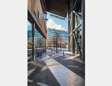 Straßenansicht im Sony Center in Berlin mit modernen Glas- und Stahlkonstruktionen, Gehwegplatten mit taktilen Markierungen und einem Starbucks-Café auf der linken Seite. Im Hintergrund sind Gebäudefassaden mit großen Fenstern und kahle Bäume zu sehen, bei sonnigem Wetter.