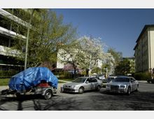 Straßenszene in einem Wohngebiet mit blühenden Bäumen im Frühling, geparkten Autos und einem Anhänger mit einer blauen Plane bedeckt. Im Hintergrund sind mehrstöckige Wohngebäude und klare blaue Himmel zu sehen.