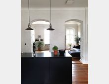 Moderne Wohnküche mit Blick auf einen offenen Wohnbereich. Helle Ziegelwände mit Rundbögen, ein schwarzer Küchenblock im Vordergrund, Hängelampen und Holzboden; im Hintergrund Pflanzen, ein Sofa und große Fenster.