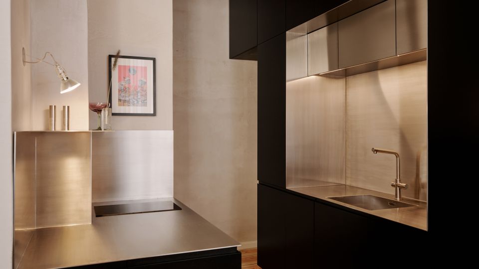 Moderne Küche mit matt schwarzen Schrankfronten und einer durchgehenden Edelstahl-Arbeitsfläche samt Spüle, Rückwand und Oberschränken. Links steht ein Edelstahlblock mit Wandlampe und gerahmtem Bild vor hellen, glatten Wänden; der Boden besteht aus Holzdielen.