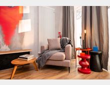 Gemütliche Wohnecke mit beigem Sessel und grauer Decke neben einem großen Fenster mit langen grauen Vorhängen. Davor stehen ein niedriger Holztisch mit Büchern sowie farbige Beistelltische mit Lampen; an der Wand befinden sich ein weißer Heizkörper und ein abstrakter roter Wandbereich.