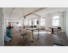 Ein heller Raum mit Holzboden, weißen Wänden und mehreren großen Fenstern, durch die Tageslicht einfällt. Der Raum ist spärlich möbliert mit unterschiedlichen Stühlen, Tischen und einem mobilen Heizkörper, die eine einfache, industriell angehauchte Atmosphäre schaffen.