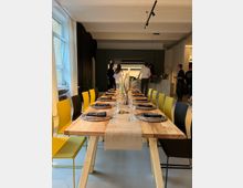 Ein stilvoll gedeckter Tisch aus Holz mit gelben und schwarzen Stühlen in einem modernen, offenen Raum mit einer Küchenzeile im Hintergrund. Auf dem Tisch befinden sich Gläser, braune Platzsets und Kerzen, während im Hintergrund Menschen in der Küche stehen.